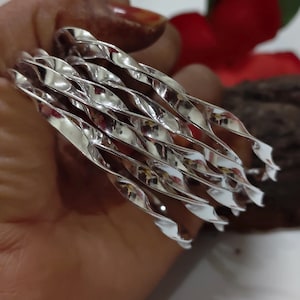Può includere: Un set di braccialetti in metallo argentato attorcigliato. I braccialetti presentano un design a spirale e riflettono la luce, creando un aspetto lucido. I gioielli sono presentati su uno sfondo sfocato.