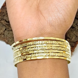 Könnte beinhalten: Ein Stapel goldfarbener Armreifen an einem Handgelenk. Die Armreifen haben ein Blumenmuster und eine gehämmerten Textur. Der Schmuck wird am Arm einer Person präsentiert.