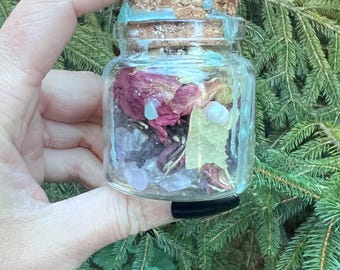 May include: A small, clear glass jar filled with dried flowers, crystals, and other natural elements. The jar has a cork stopper with blue accents. The background is a backdrop of green pine needles and foliage.