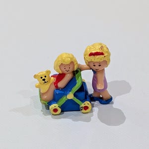 May include: A miniature toy scene featuring two figures and a stroller. One figure is pushing the stroller, while the other figure is seated inside. A small teddy bear is also in the stroller. The figures have blonde hair and are wearing colorful clothing.
