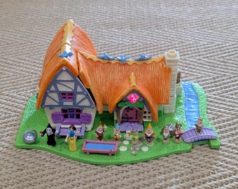 May include: A miniature cottage toy with an orange thatched roof, a white chimney, and a green base. The scene includes miniature figures of Snow White, the Evil Queen, and the Seven Dwarfs. A small stream and bridge are also present.