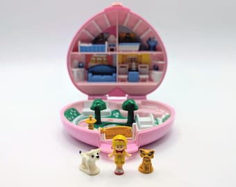 May include: A heart-shaped pink plastic playset that opens to reveal a two-story interior with furniture and a garden area. Included are three small figurines: a yellow-clad character, a white dog, and a brown cat, all positioned in front.