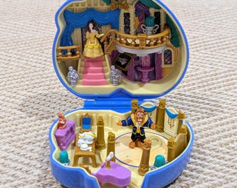 Puede incluir: Un juego de juguetes azul en forma de concha con figuras y muebles en miniatura. El nivel superior tiene una figura con un vestido amarillo en una escalera rosa. El nivel inferior tiene una figura de la bestia con muebles. El juguete está abierto.