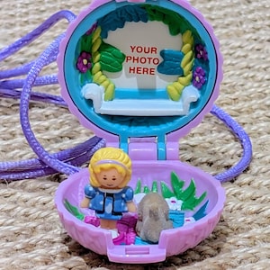 May include: A vintage, open, purple locket with a miniature doll and a small animal inside. The locket has a white bench and the text "YOUR PHOTO HERE". A purple cord is attached to the locket.