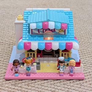 May include: A miniature pizza restaurant playset with a blue roof, pink and white striped awning, and four small figures. The building has a pink brick base and a sign that says "Pizza".