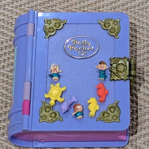 May include: A vintage Polly Pocket playset in a book-like design. The purple exterior features gold-colored accents and a clasp. Inside, miniature figures and sea creatures are arranged on a blue surface, with the Polly Pocket logo visible.