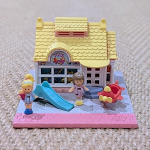 May include: A miniature toy shop with a yellow roof and the word "TOYS" above the window. Includes a blue slide, two small figures, and a red aeroplane. The base is pink with a brick pattern.