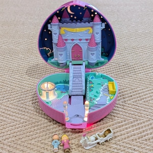 May include: A pink heart-shaped toy castle playset, open to reveal a miniature castle with pink turrets, a bridge, and a small garden. Includes miniature figures and a horse-drawn carriage. The background features a night sky with a crescent moon and stars.