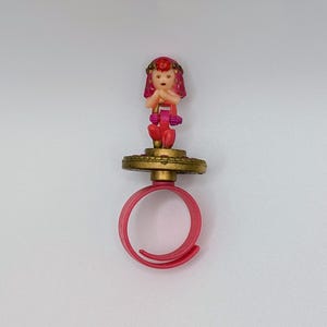 May include: A pink plastic ring featuring a small figurine of a person with pink hair and a red gem on their forehead. The figurine stands on a circular, textured gold-colored base that is attached to the adjustable pink band of the ring.