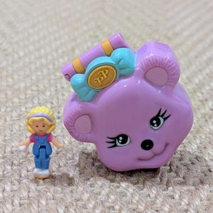 May include: A vintage, lilac-coloured toy shaped like a bear's head with a candy-shaped bow. A small doll with blonde hair and blue dungarees stands nearby. The bow has the letters "BP" on it.