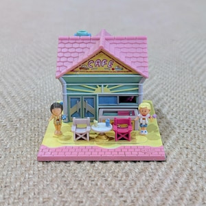 May include: A miniature pink cafe playset with a pink roof and the word "CAFE" above the entrance. The cafe has two small chairs, a table, and two miniature figures. The base is yellow and pink.