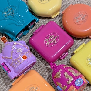 May include: A collection of Polly Pocket plastic cases in various shapes and colours, including teal, yellow, orange, magenta, and purple. Some cases are simple geometric shapes, while others feature ornate, detailed designs with embossed text.
