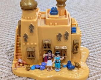 May include: A tan plastic toy castle with gold accents, featuring two figures and a monkey. The castle has two gold-topped towers, windows, and a staircase. The figures are dressed in blue and white outfits. The toy is set on a tan base.