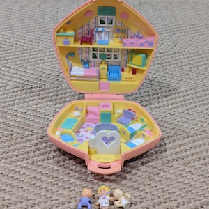 May include: A vintage, open, pink and yellow dollhouse with multiple rooms, furnished with miniature beds, chairs, and other accessories. Three small dolls are in front of the dollhouse. The dollhouse is on a textured, tan surface.
