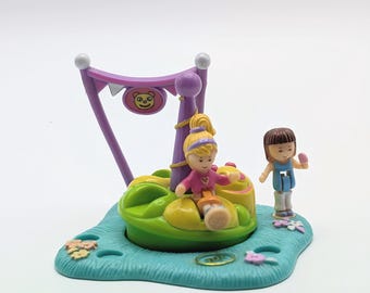 Könnte beinhalten: Ein Spielzeug-Set aus Kunststoff mit einer grünen Karussellbasis mit gelben Sitzen, einem violetten Stützrahmen mit einem Bärengesicht-Schild und zwei kleinen Figuren. Eine blonde Figur sitzt auf dem Karussell, während eine zweite Figur daneben steht.