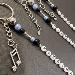 May include: Keychains with silver-tone chains, beads, and musical note charms. Some keychains have letter beads spelling "POKOTHO". The beads are black, light blue, and silver. The musical note charms have small crystals.