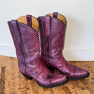 May include: A pair of tall, purple cowboy boots with a snakeskin pattern. The boots have a pointed toe, a stacked heel, and a pull-on design. The interior is a light tan colour.