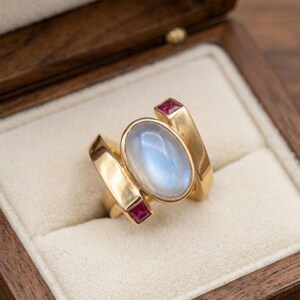 May include: A gold ring with an oval, light blue moonstone center and two small, square red gemstones. The ring has a unique, modern design and is displayed in a wooden jewelry box.
