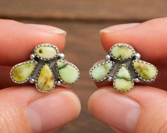 Op de afbeelding: Een paar zilverkleurige oorstekers, elk met een cluster van lichtgroene en gele edelstenen. De stenen zijn druppel- en ovaalvormig, gezet in een geschulpte bezel. De oorbellen worden door vingers vastgehouden.