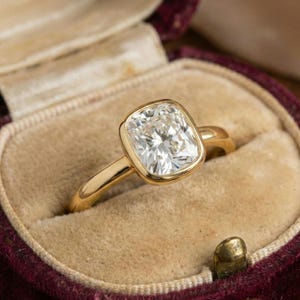 Peut inclure: Une bague en or avec un gros diamant de taille carrée serti dans une lunette. Le diamant clair et étincelant est présenté dans un écrin en velours bordeaux.