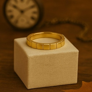 Puede incluir: Un anillo dorado con un patrón cuadrado descansa sobre una caja beige. Un reloj de bolsillo borroso está en el fondo. El ambiente cálido incluye una hoja, creando un ambiente clásico y otoñal.