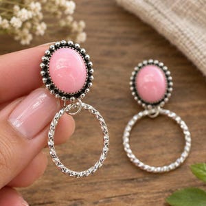 Può includere: Un paio di orecchini con una pietra rosa liscia di forma ovale incastonata in una cornice argentata con bordo a perline. Un cerchio in metallo intrecciato color argento pende dalla base di ogni montatura ovale, creando un design pendente.