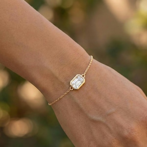 Peut inclure: Un bracelet en or orné d'une pierre précieuse rectangulaire taille émeraude. La pierre est sertie dans une lunette en or et reliée à une fine chaîne en or. Le bracelet est porté sur un poignet.