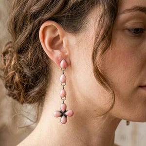May include: A long drop earring featuring five smooth, pale pink, teardrop-shaped stones set in silver. The stones are arranged vertically in a line, with the bottom four stones forming a cross-like shape at the base of the earring.