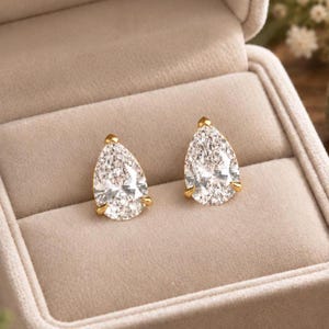 May include: A pair of stud earrings featuring pear-shaped clear stones set in gold-toned metal prongs. The earrings are displayed side by side, showcasing the faceted surfaces of the stones and the metallic settings that secure each gem in place.