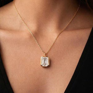 Könnte beinhalten: Eine goldene Halskette mit einem rechteckigen, im Smaragdschliff facettierten, klaren Edelsteinanhänger in einer Goldfassung. Der Anhänger hängt mittig an einer feinen Goldgliederkette und liegt auf dem Schlüsselbeinbereich mit poliertem Metallglanz.