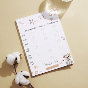 Può includere: Un blocco note bianco con il testo "Meine Tricks" e un elenco di comandi di addestramento per cani. Include un'illustrazione di cucciolo di cartone animato e impronte di zampe. Un bicchiere d'acqua e steli di cotone sono anche nell'immagine.