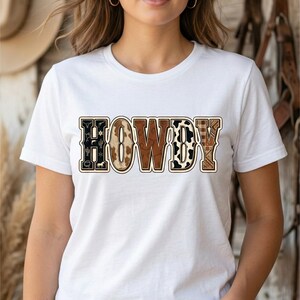 Puede incluir: Camiseta blanca con la palabra "HOWDY" en un diseño de temática western. Las letras presentan patrones como piel de vaca, cuadros y detalles florales. La camiseta es de estilo cuello redondo clásico.