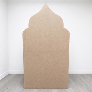 May include: A large, freestanding, unfinished wooden archway with a decorative top. The archway is made of light brown wood and stands upright against a white wall and grey floor.
