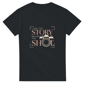 Könnte beinhalten: Ein schwarzes T-Shirt mit kurzen Ärmeln und einem Grafikdesign in Hellrosa und Weiß. Das Design zeigt eine Kamera-Illustration und den Text I SEE THE STORY BEHIND THE SHOT, der in einem rechteckigen Rahmen auf der Brust platziert ist.