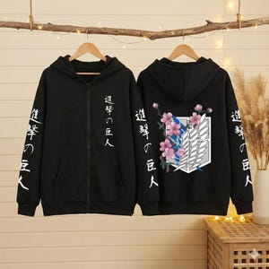 May include: Black zip-up hoodie with white Japanese text on the sleeves and down the front. The back of the hoodie features a white and gray emblem with pink flowers and blue accents. The hoodie is hanging on a wooden hanger.