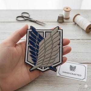 May include: Embroidered patch with a shield design and blue and beige wings. The patch has a black and white border. A small white card with the text "HANDCRAFTED" is also visible.