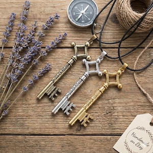 Può includere: Tre ciondoli a forma di chiave in oro, argento e bronzo, ciascuno con testo giapponese, sono esposti su una superficie di legno. Sono presenti anche una bussola, spago e rametti di lavanda. I ciondoli sono su cordini neri.