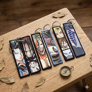 Puede incluir: Llaveros bordados con personajes de anime y anillos plateados. Los llaveros rectangulares tienen diseños coloridos y bordes negros. También se ve una pequeña brújula sobre una superficie de madera.