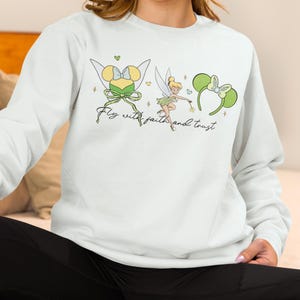 Puede incluir: Una sudadera blanca con un gráfico de Campanilla y diademas de orejas de ratón. El diseño incluye el texto "Fly with faith and trust" en una fuente cursiva. El gráfico es en tonos de verde, amarillo y negro.