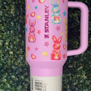 May include: A pink Stanley Quencher Protour tumbler with a handle, decorated with colorful bunny illustrations, hearts, and stars. The tumbler holds 40 oz (1.18 L) and features the Stanley logo.