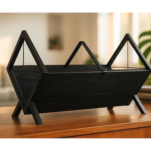 May include: A black wooden magazine rack with a geometric design. The rack features a rectangular body supported by angled legs and topped with three triangular handles. The wood grain is visible, and the rack sits on a wooden surface.