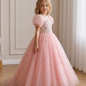 May include: A floor-length pink dress featuring a bodice covered in shimmering sequins and beads, short puffed sleeves, and a full tulle skirt adorned with scattered silver glitter stars. The dress has a round neckline and a pearl-embellished headband.