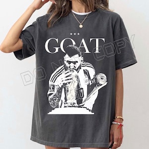 Puede incluir: Camiseta gris oscuro con un gr&aacute;fico en blanco y negro de un jugador de f&uacute;tbol sosteniendo un trofeo. La palabra "GOAT" est&aacute; impresa encima de la imagen, con tres estrellas. La camiseta es de estilo casual.