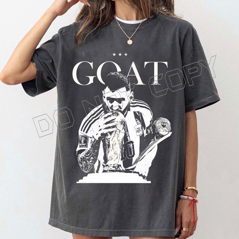 Puede incluir: Camiseta gris oscuro con un gr&aacute;fico en blanco y negro de un jugador de f&uacute;tbol sosteniendo un trofeo. La palabra "GOAT" est&aacute; impresa encima de la imagen, con tres estrellas. La camiseta es de estilo casual.