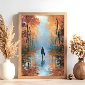 Könnte beinhalten: Ein gerahmter Kunstdruck, der eine Person zeigt, die durch einen herbstlichen Wald mit orangefarbenen und roten Bäumen geht, gespiegelt in einer Pfütze. Das Bild wird von einer weißen Vase mit Gräsern und einer braunen Vase mit Blumen flankiert.
