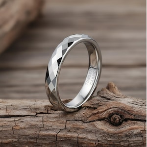 May include: A silver tungsten carbide ring with a geometric, faceted design. The ring is shown on a piece of weathered wood. The text "TUNGSTEN CARBIDE" is visible on the inside of the ring.