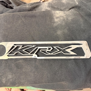 May include: A silver metal plate with the word "KRX" cut out in a bold, geometric font. The plate has a circular hole on the left side and two smaller holes on the right side. The plate is rectangular and sits on a dark gray surface.