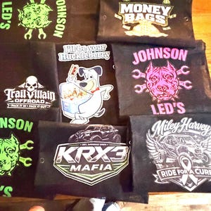 May include: A collection of black fabric pouches with various graphic designs and text. Designs include a skull, a dog, a car, and the words "Money Bags," "KRX3 Mafia," and "Ride for a Cure." Some pouches have the name "Johnson."