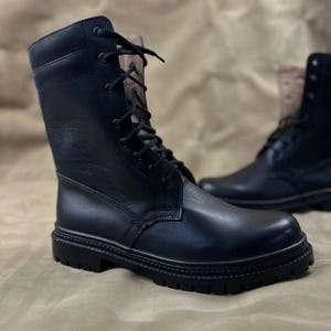 May include: Black leather combat boots with a lace-up design. The boots have a rounded toe and a thick, lugged sole. The shaft of the boot extends up the ankle. The background is a neutral tan colour.