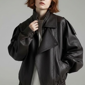 Peut inclure: Veste en cuir marron foncé avec une finition texturée. La veste présente un large col, une fermeture éclair diagonale et une taille ceinturée. Les manches sont longues et légèrement bouffantes aux épaules. Vêtement d'extérieur élégant.